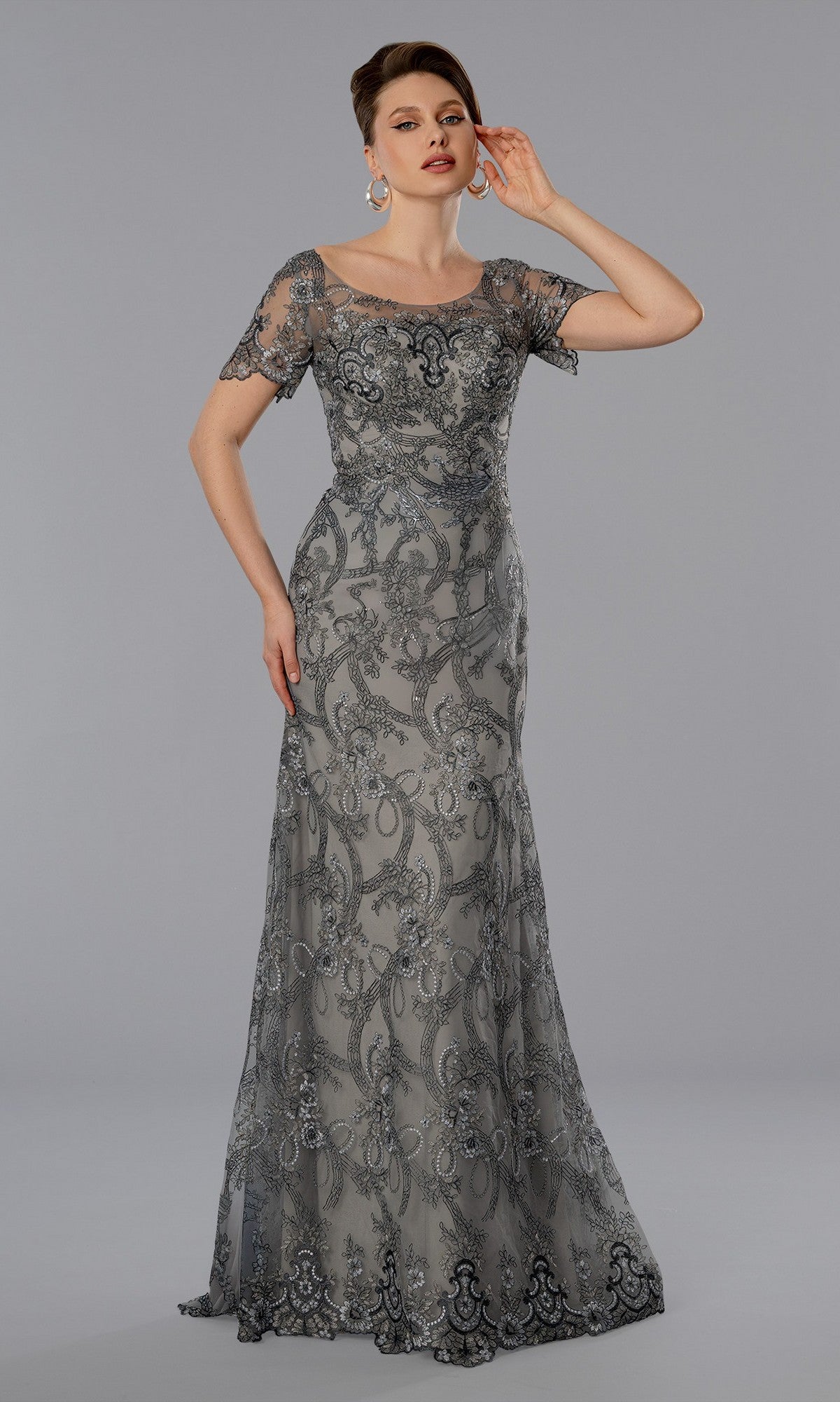 A woman in the Stella Couture 25369 long formal dress, gray with sheer short sleeves and intricate embroidery, stands against a plain gray background.